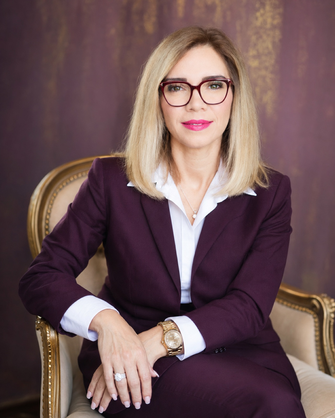 Plum suit, burgundy frames. Seated in a carved gilt armchair against a plum wall.