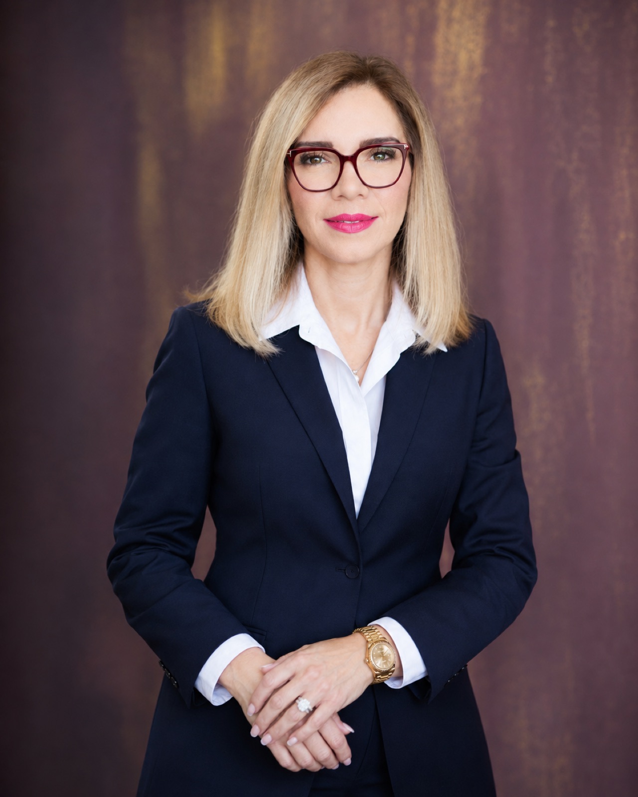 Navy suit, tortoiseshell frames. Hands folded, bronze studio ground.