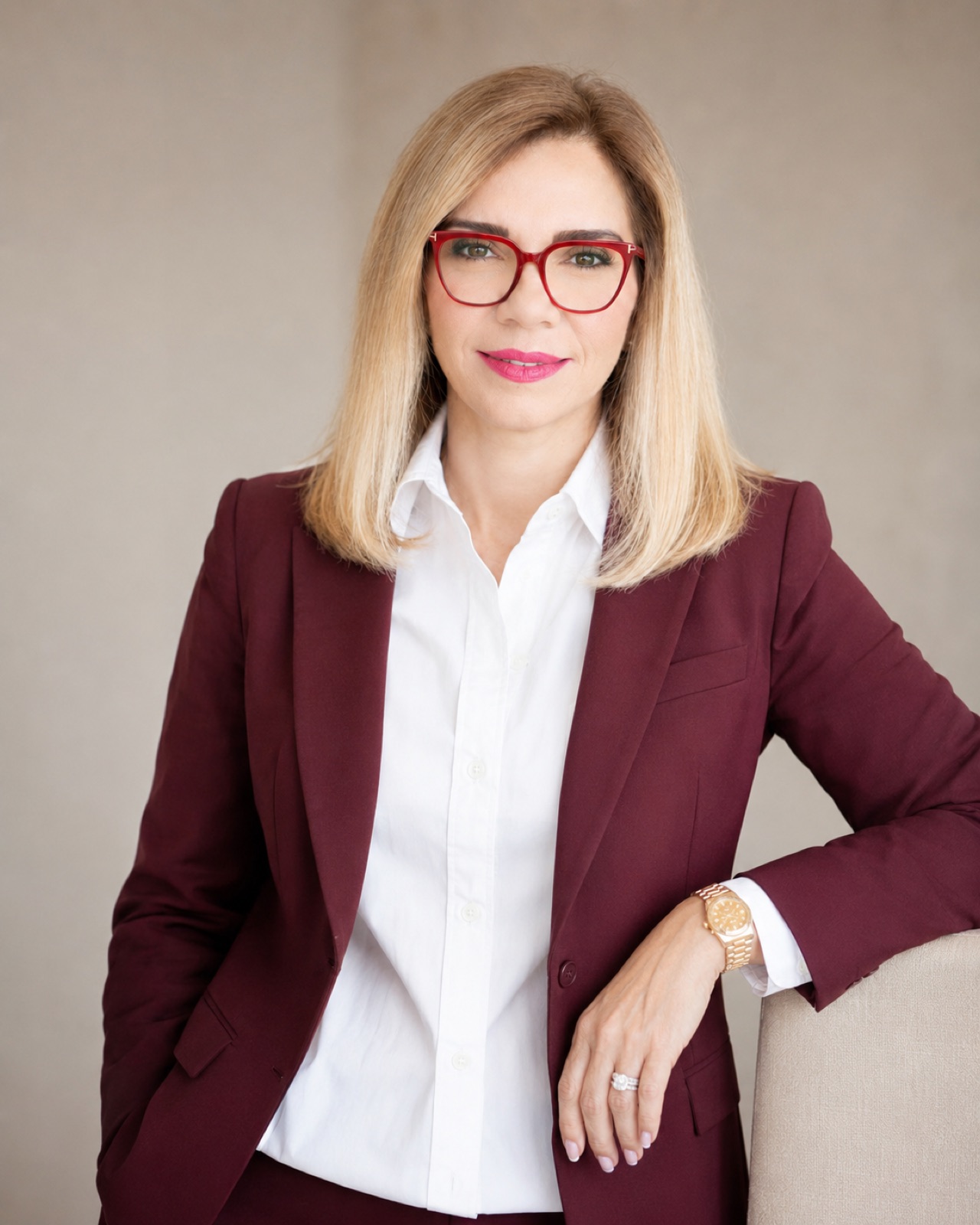 Burgundy suit, red frames. Standing, arm along an armchair back.
