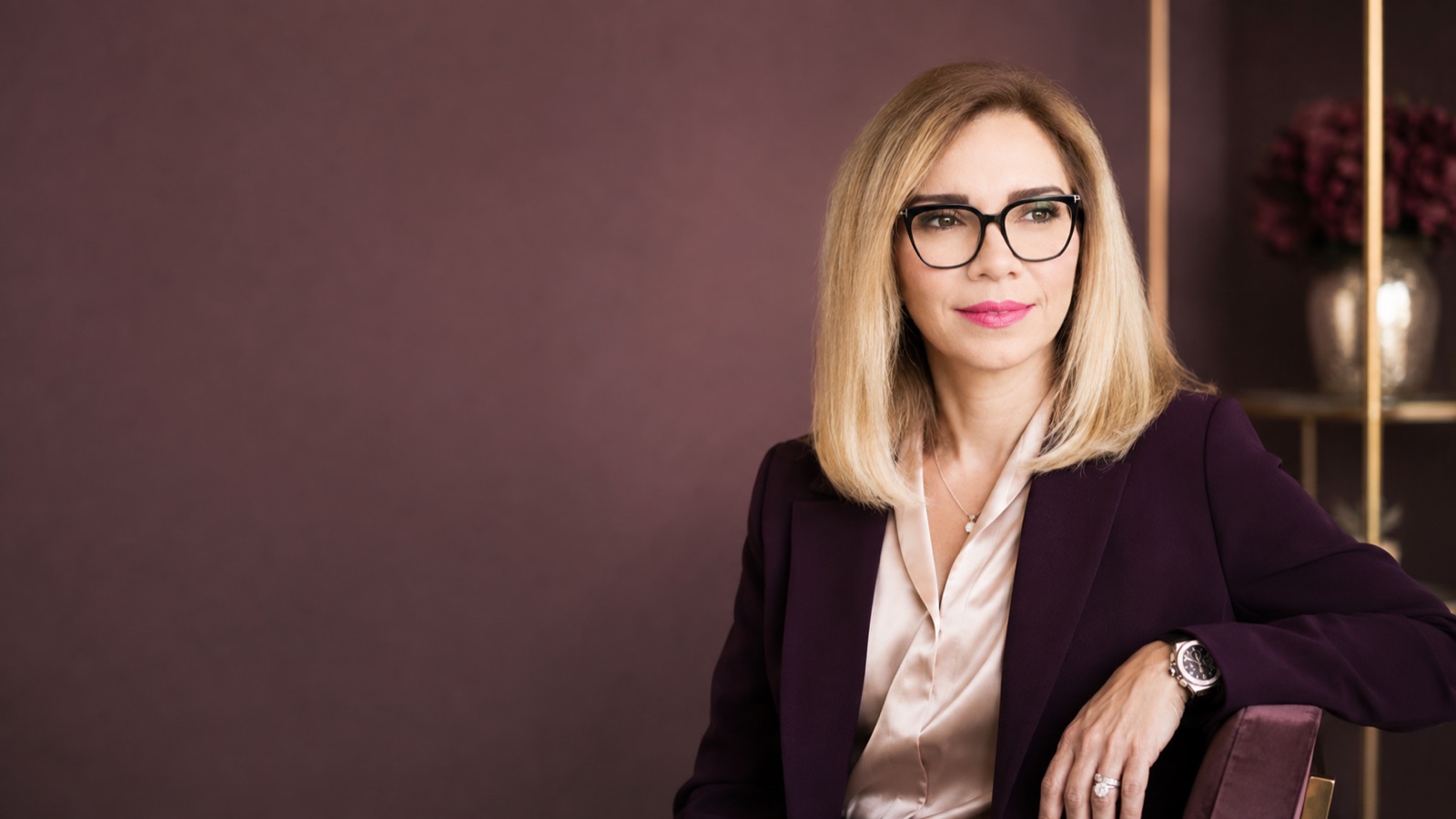 Plum blazer over a champagne blouse, black frames. Plum wall, subject right — clean negative space at left for a headline.