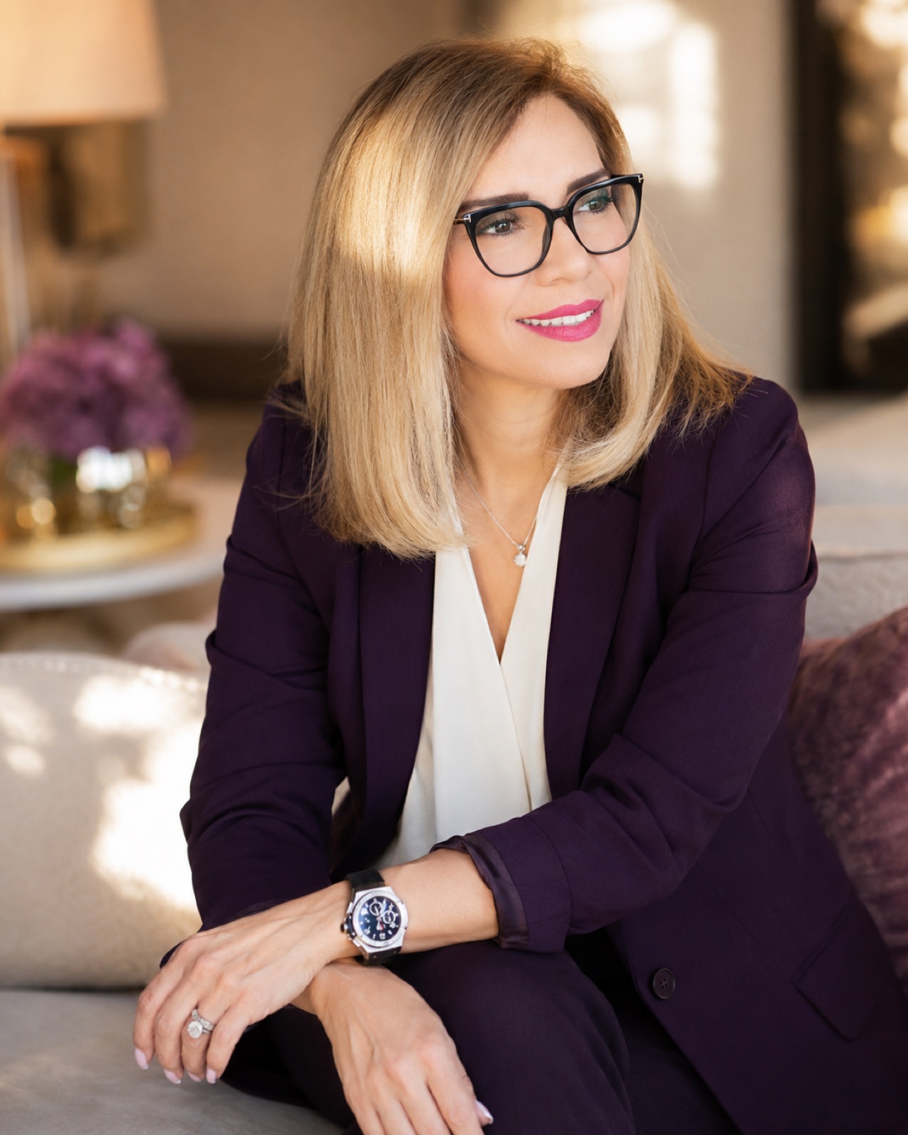 Plum suit over a cream blouse, black frames. Warm lamplit lounge.