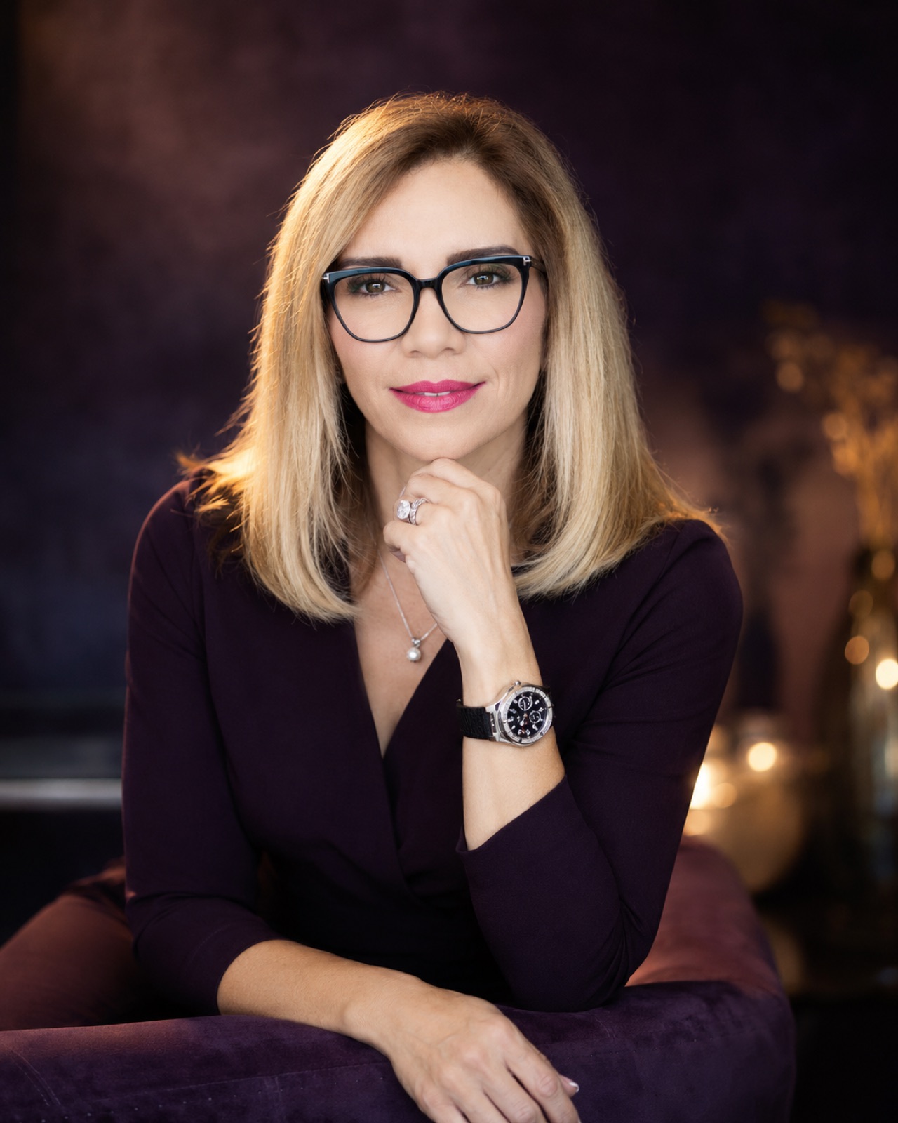 Plum knit, black frames. Hand to chin, candlelit plum ground. The darkest, most intimate grade.