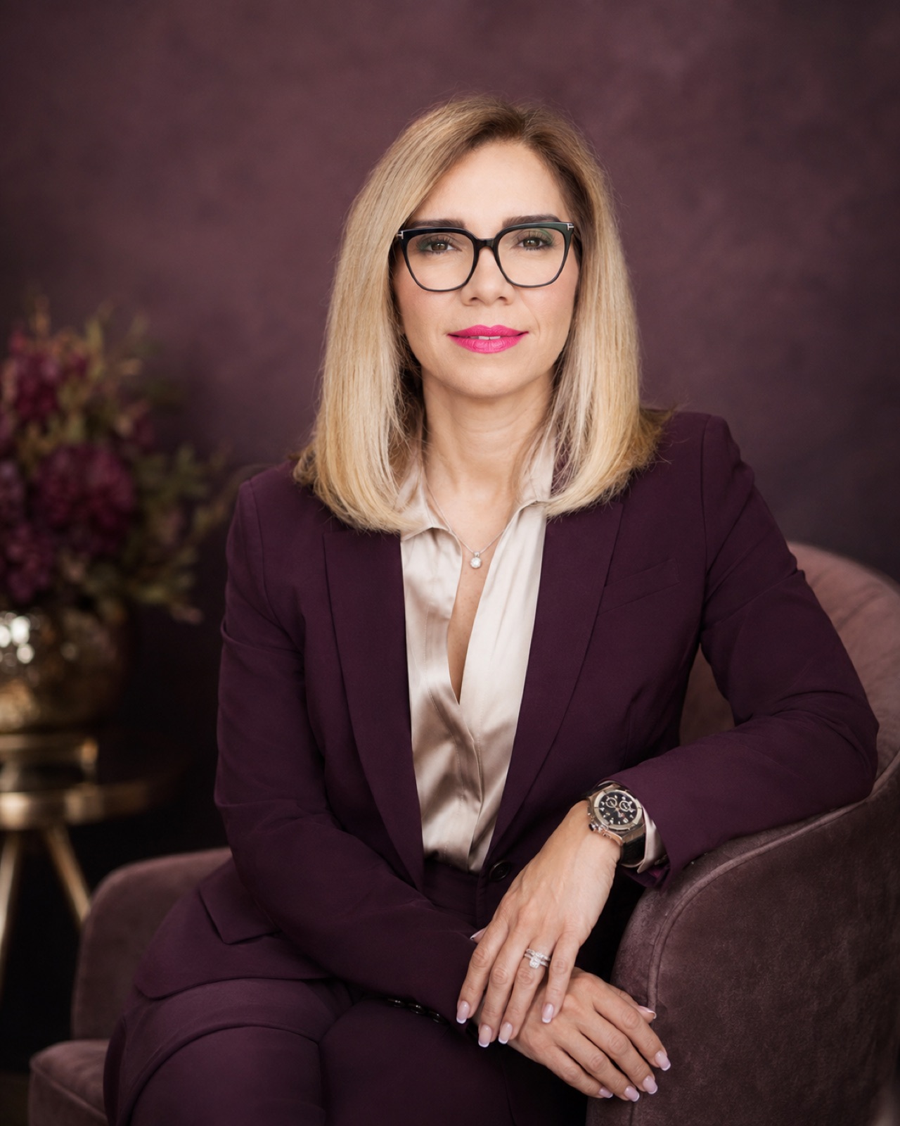 Plum suit over a champagne blouse, black frames. Seated, plum ground.