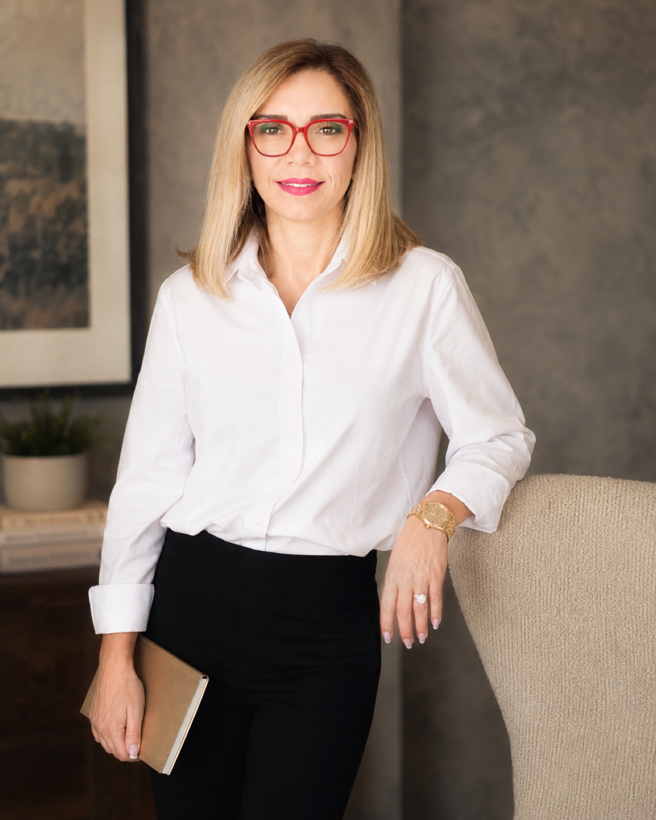 White shirt over black trousers, red frames. Standing with a tan notebook, hand resting on an armchair back. Crops mid-thigh.