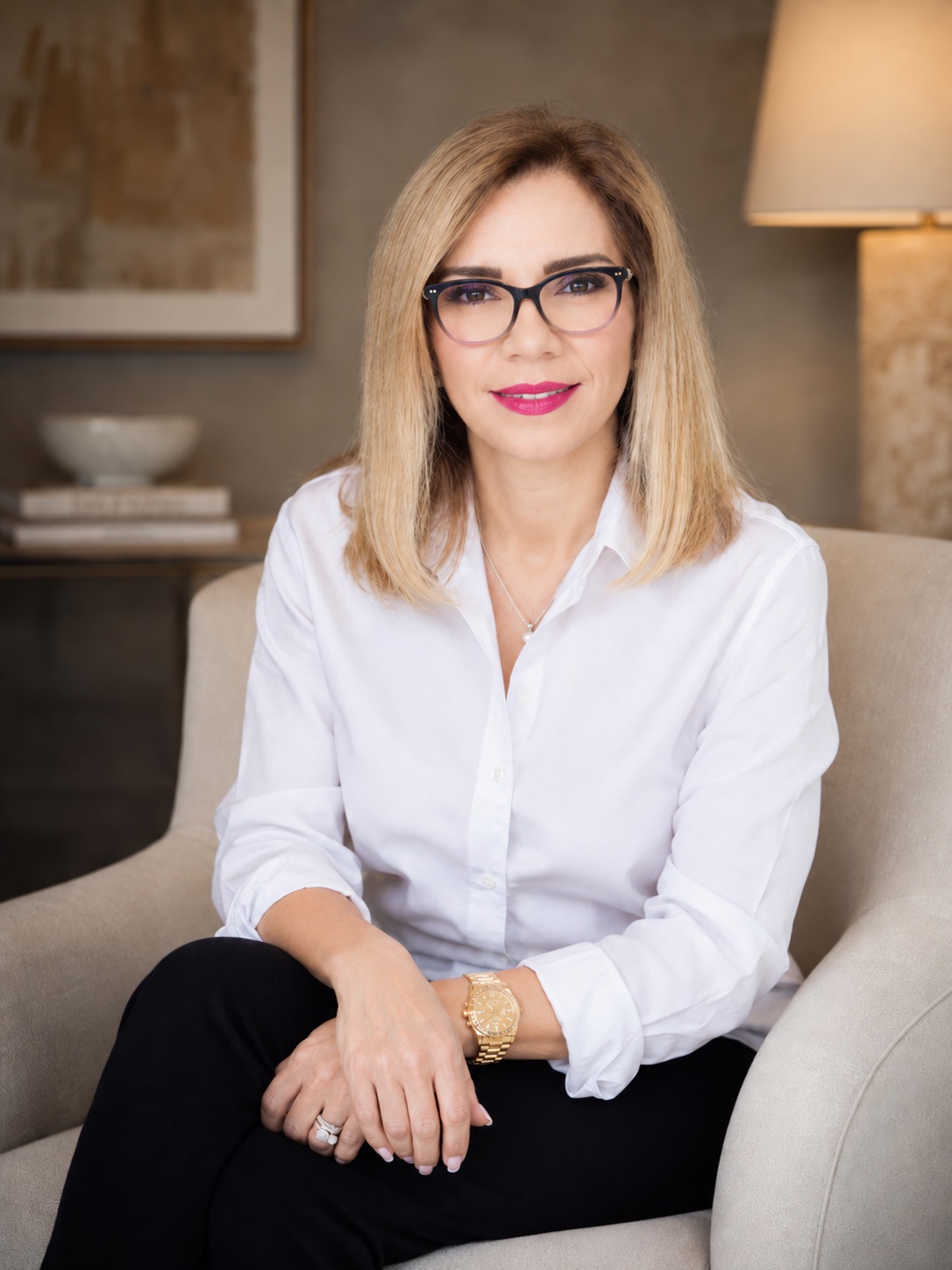 Black frames. Seated in a cream armchair, hands on the knee, lamp and artwork behind. Same setup as 65.